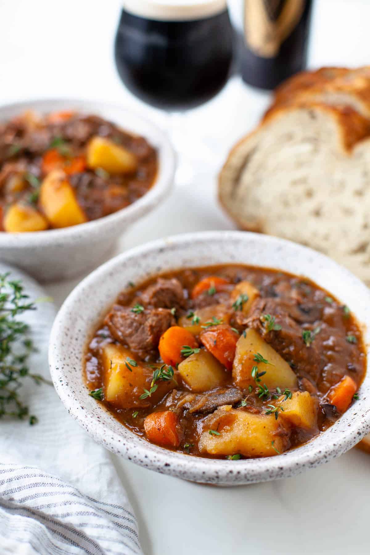 Bowl of instant pot guiness beef stew sits close by. Glass and can of guiness can be seen in the distance. Loaf of bread sits to the right. Another pot of stew and a blue and white striped towel sits on the left